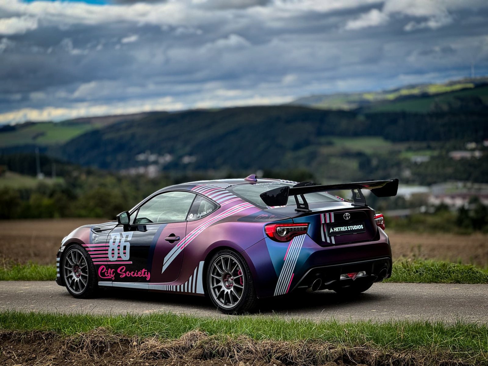 Folierter Toyota GT86 in Pink, Violett und Blau mit Hetrex Studios Branding und großem Heckspoiler auf einer Landstraße vor einer hügeligen Landschaft.
