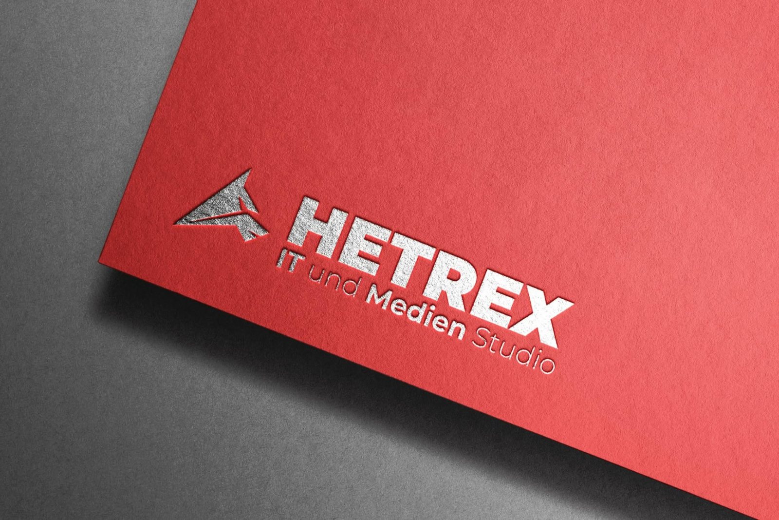 Nahaufnahme des silbern geprägten Hetrex Logos mit dem Schriftzug „IT und Medien Studio“ auf rotem strukturiertem Papier.